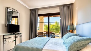 Quarto de hotel com cama, televisão em suporte, varanda com mobiliário exterior e vista para o verde.