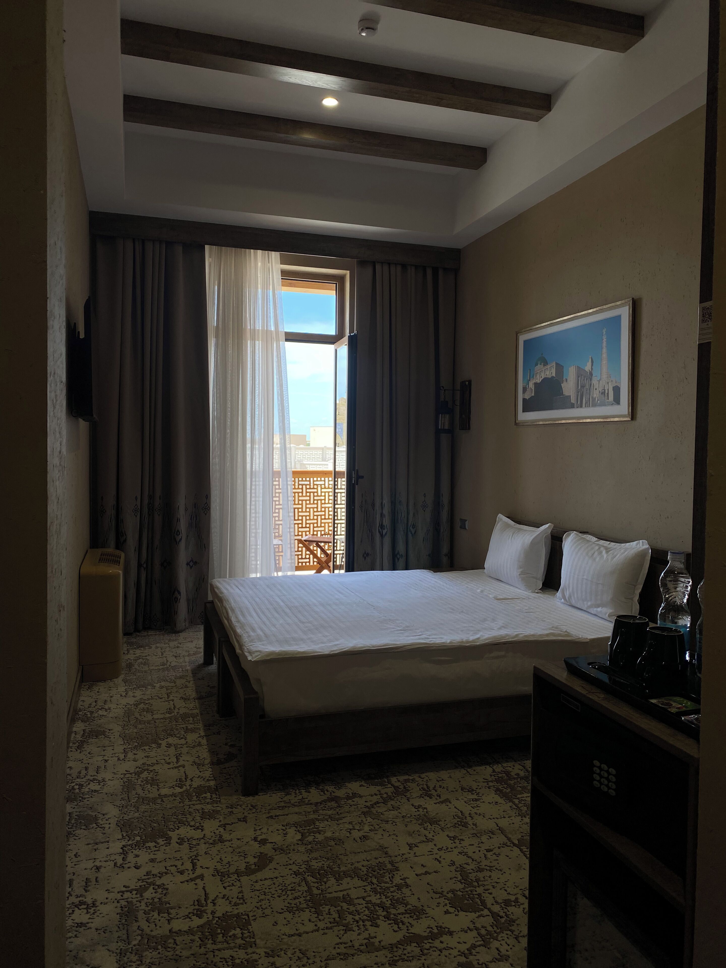 Un cuarto de hotel con una cama, un cuadro, un escritorio con un teléfono y vistas a edificios a través de la ventana.