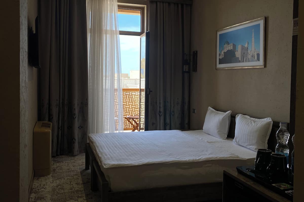 A hotel room with a bed, a painting, a desk with a phone, and a view of buildings through the window.