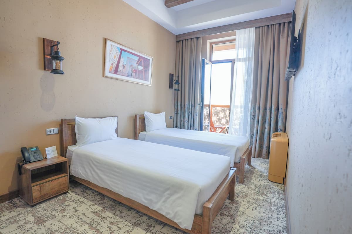 A hotel room with two beds, a wooden nightstand, a telephone, a painting on the wall, and a view of the outside through a window with curtains.