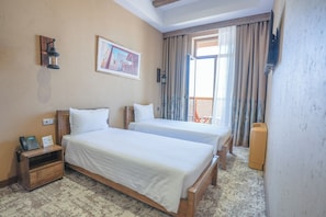 A hotel room with two beds, a wooden nightstand, a telephone, a painting on the wall, and a view of the outside through a window with curtains. - Arda Turan (Khiva)