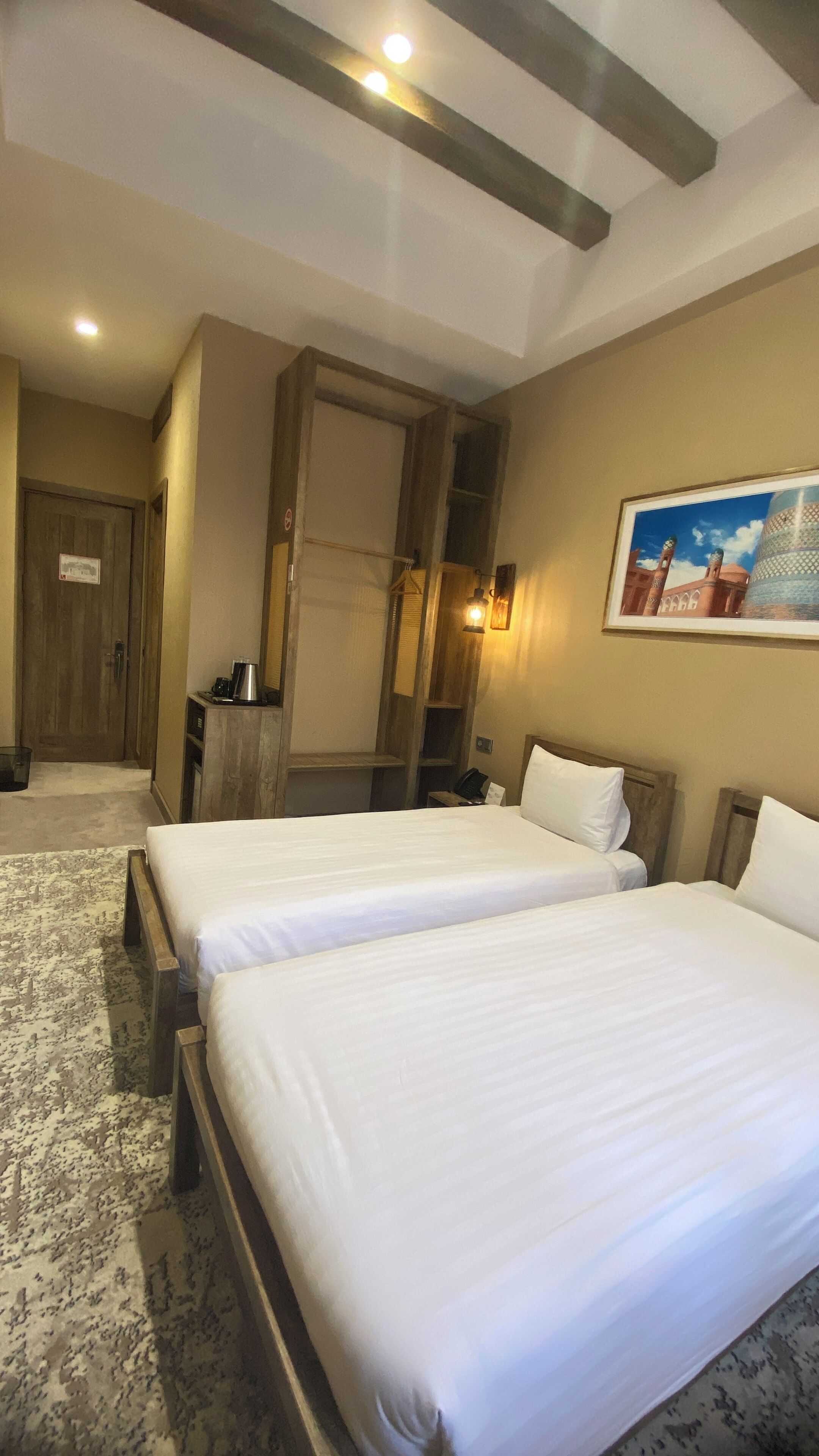Habitación de hotel con dos camas, un armario de madera, un escritorio con silla, una cafetera y un cuadro en la pared.