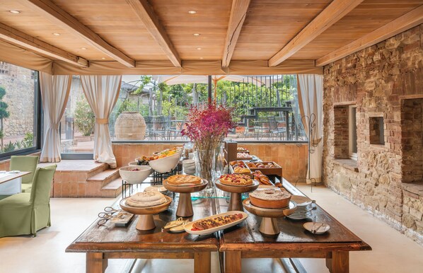 A dining area with a long wooden table set for breakfast, surrounded by stone walls and large windows. - Borgo Tre Vaselle (Torgiano)