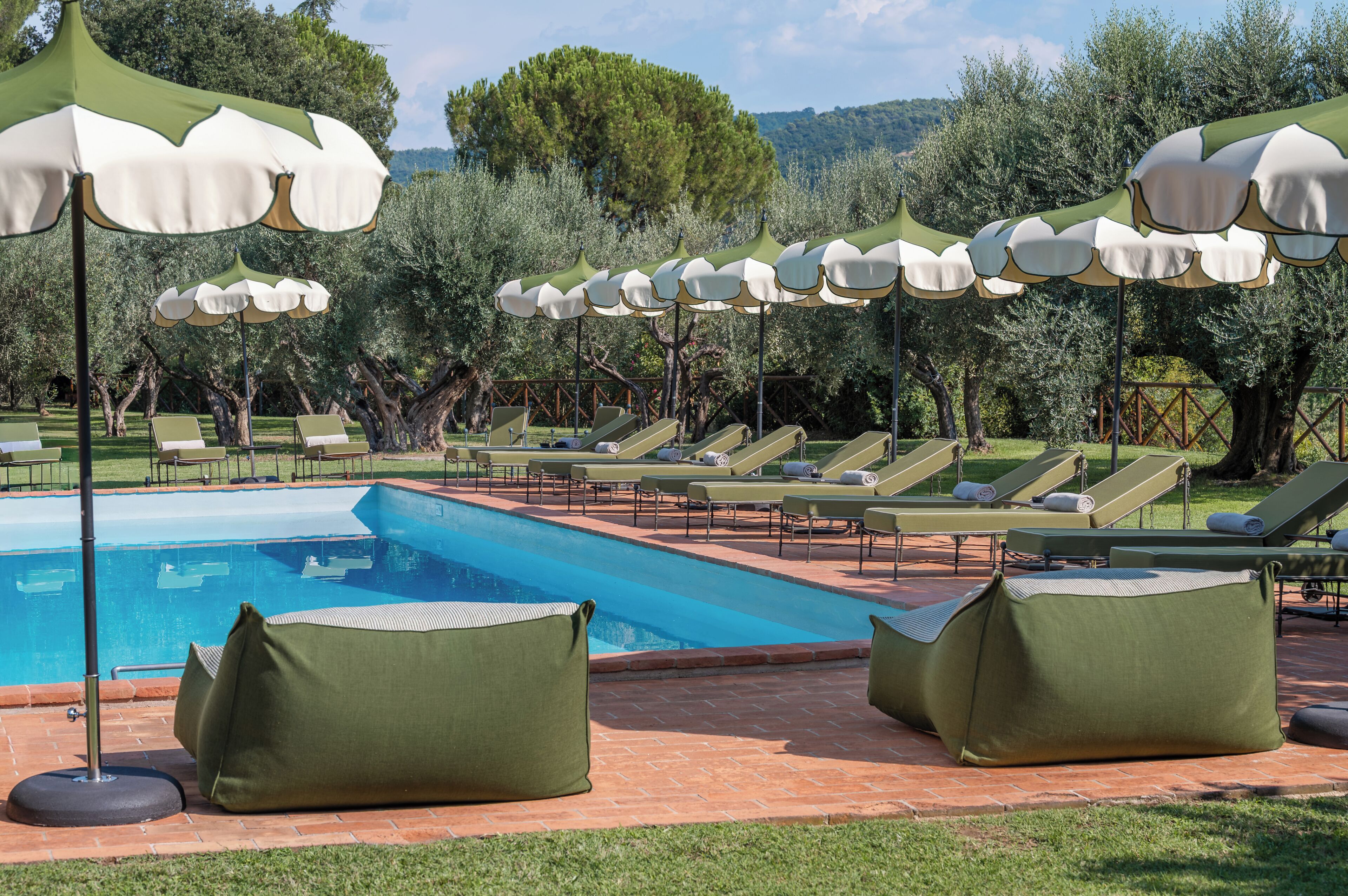 Zona junto a la piscina con sillas de descanso y sombrillas, rodeada de árboles.