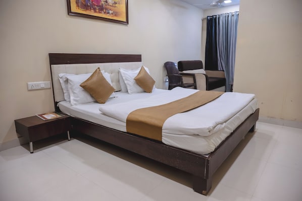A neatly made bed with white and brown bedding, a wooden headboard, a bedside table with a book, and a chair in the corner. - Park View Residency (Bhuj)