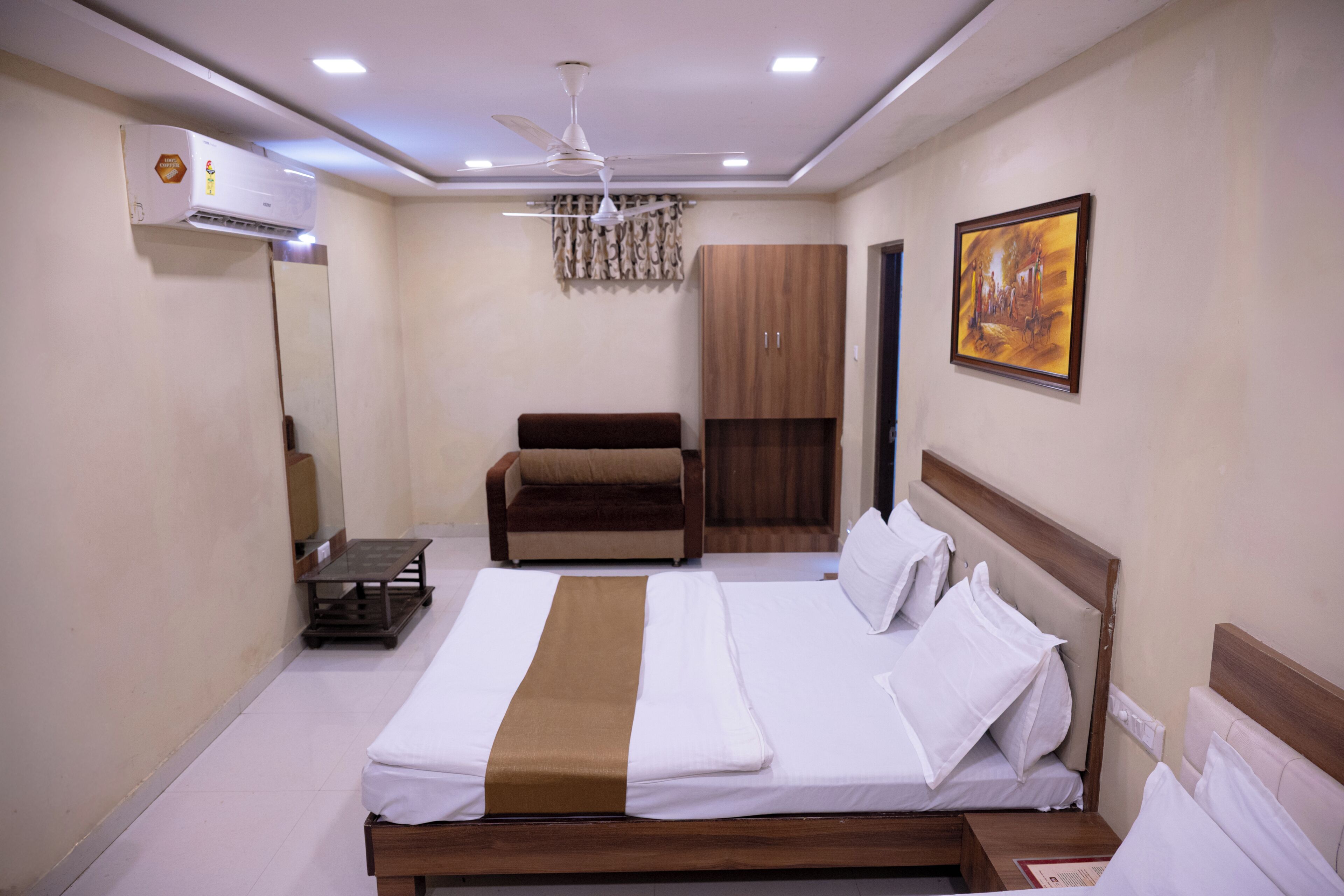 A hotel room with a bed, a sofa, a wardrobe, a ceiling fan, and a painting on the wall.