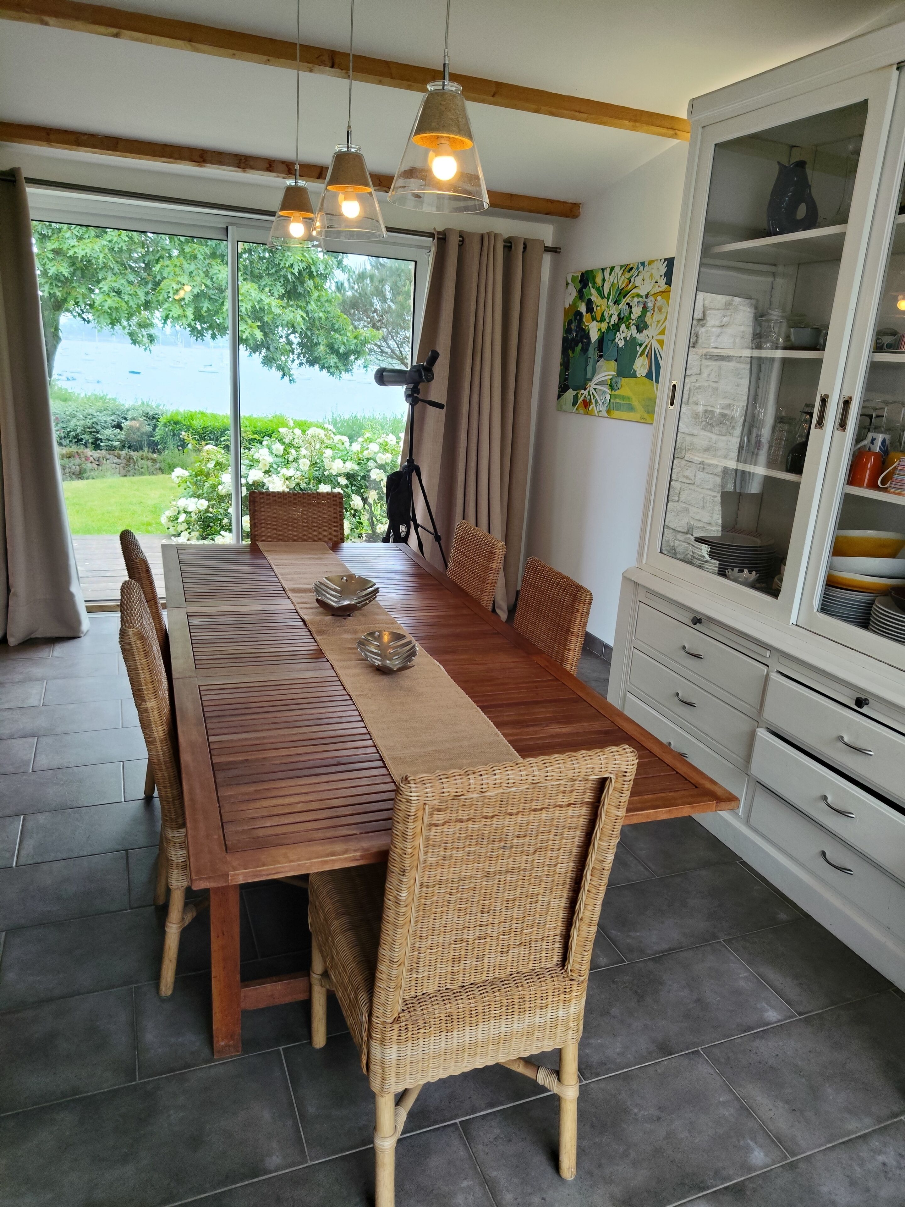 Uma área de refeições com mesa de madeira, cadeiras de vime, luminárias pendentes, um tripé e um armário com portas de vidro.