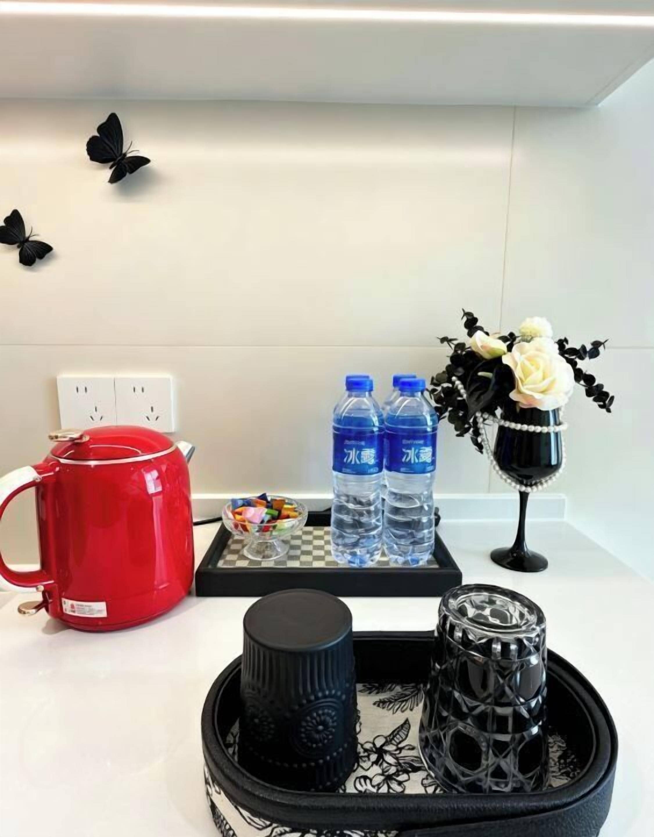 Una encimera de cocina con un hervidor rojo, dos botellas de agua, una taza negra, un vaso decorativo y un florero con flores.