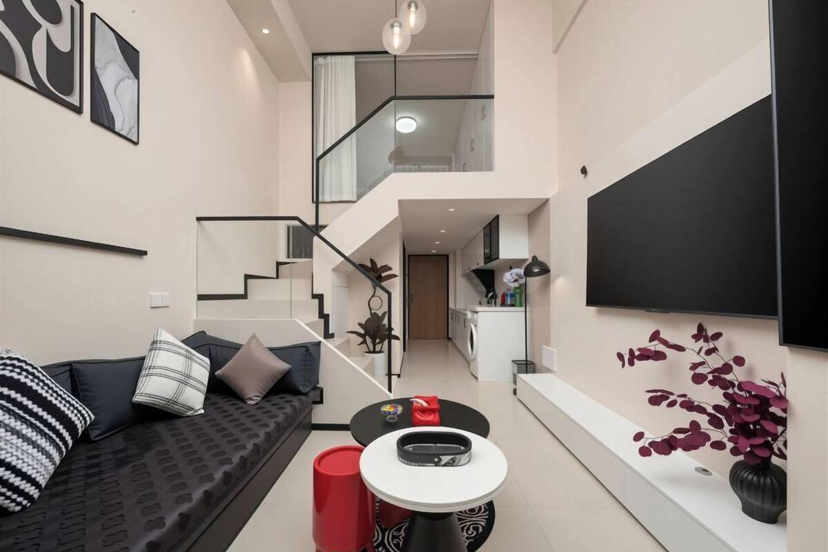 Uma sala de estar moderna com sofá, mesa de centro, um banquinho vermelho, escada, área de cozinha e uma TV de tela plana na parede.