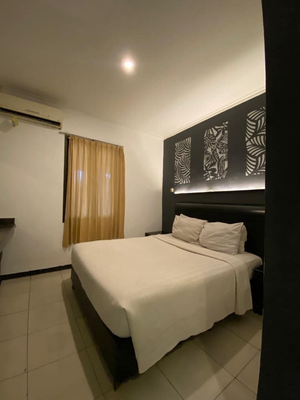 Quarto de hotel com cama, cortinas, cabeceira com painéis decorativos e luminária no teto.