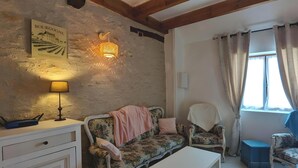 Une pièce rustique avec un mur en pierre, un canapé, un fauteuil, une petite table, une lampe et une fenêtre avec des rideaux.
