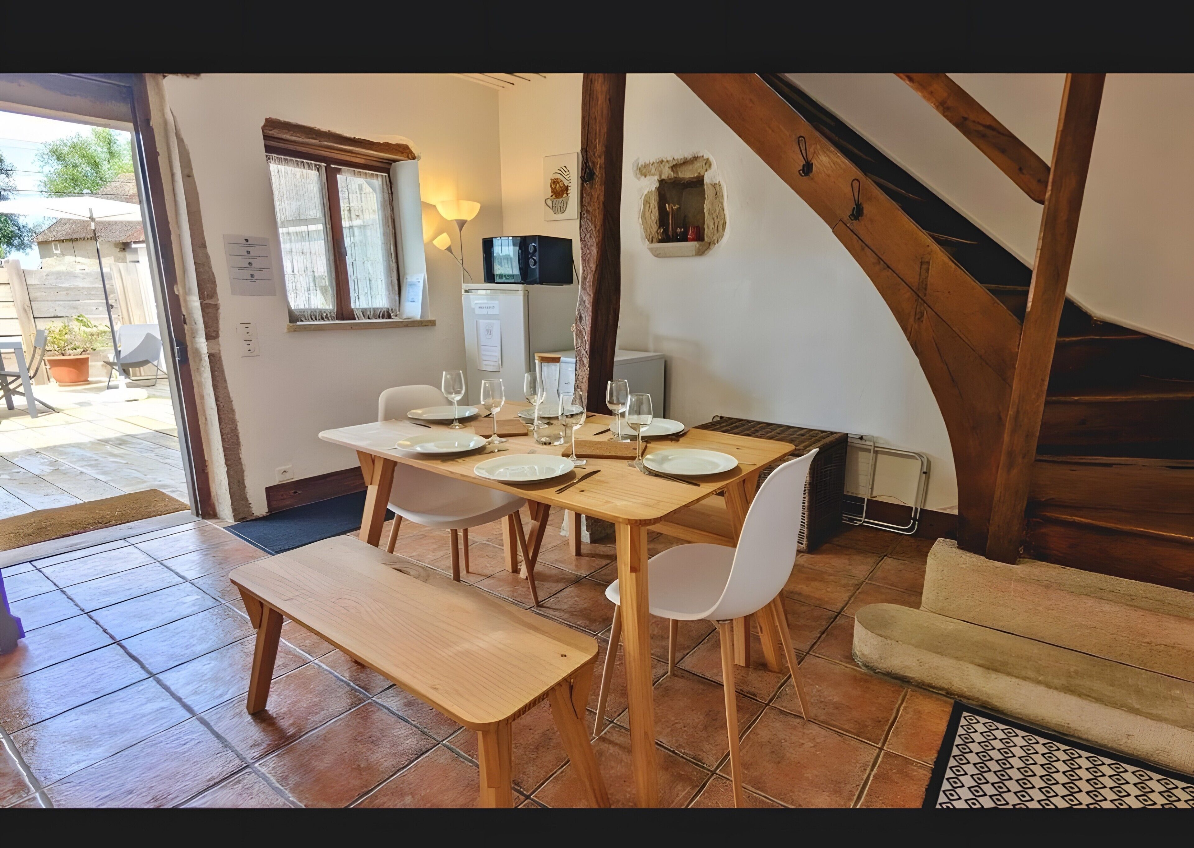 Un comedor con una mesa de madera dispuesta para cuatro, sillas blancas, un banco, un televisor pequeño y una ventana con cortinas.