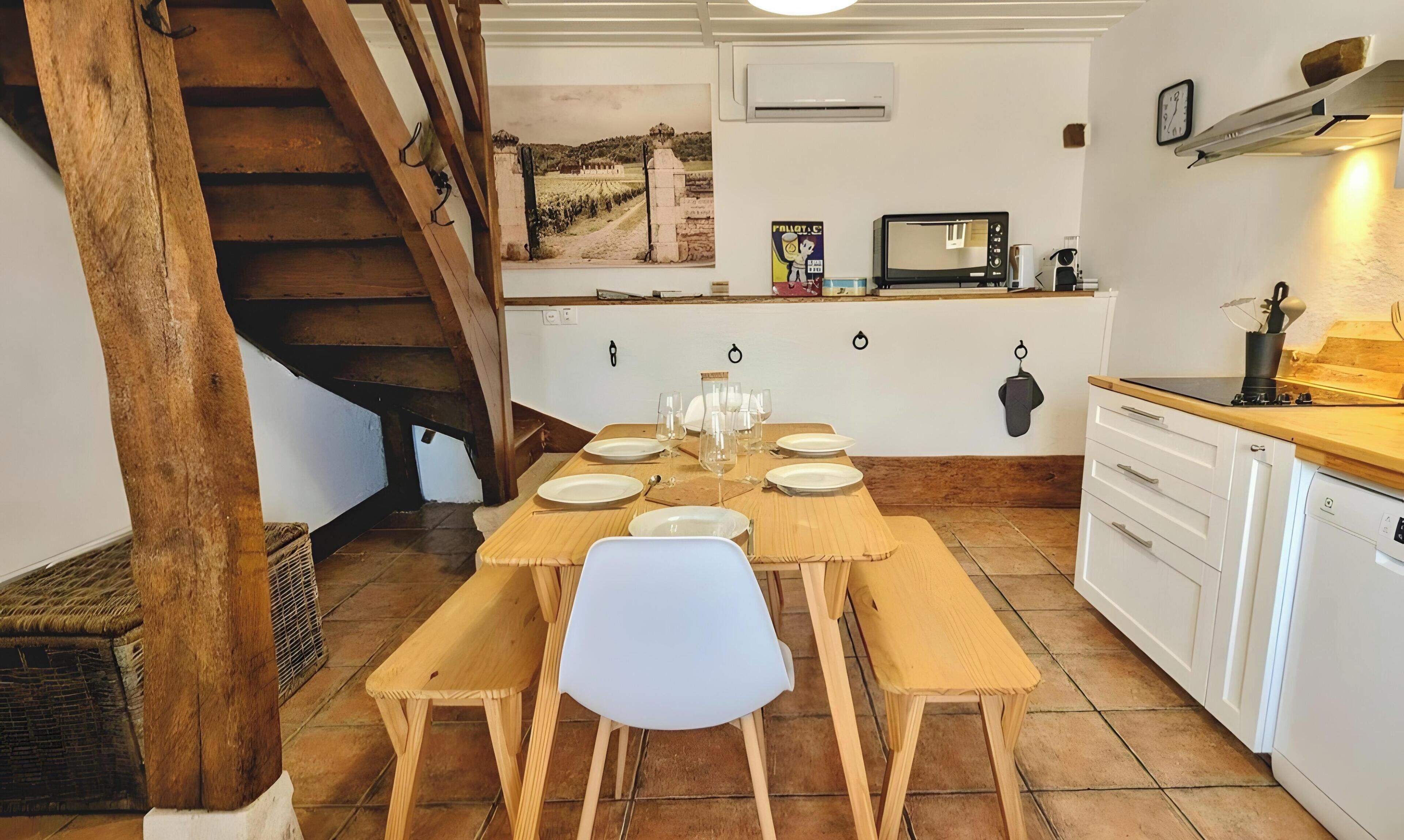 A dining area with a wooden table set for six, a white chair in the center, and a kitchen with a microwave and sink.
