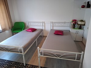 Deux lits simples avec des cadres blancs et des matelas beige, une chaise verte, une armoire blanche ornée d’un vase de fleurs et un radiateur.