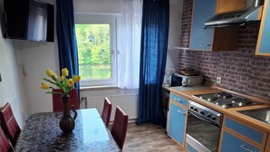 Een kleine keuken met een eettafel, stoelen, een vaas met bloemen, een raam met gordijnen en een bakstenen muur.