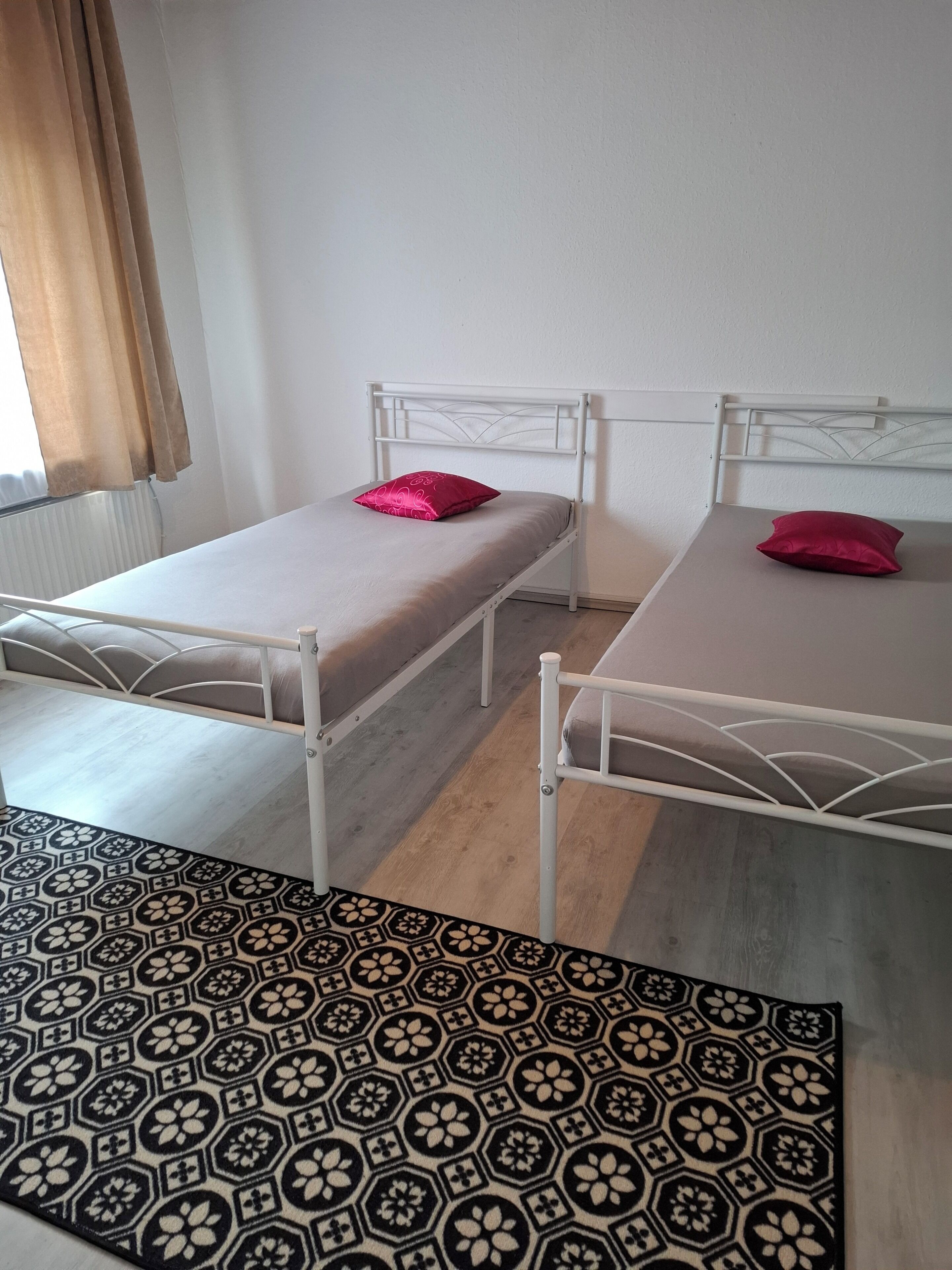 Ein Zimmer mit zwei Einzelbetten, einem gemusterten Teppich und einem Fenster mit Vorhängen.