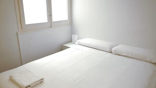 Un lit simple avec du linge de lit blanc, une fenêtre avec des stores et un mur blanc uni.