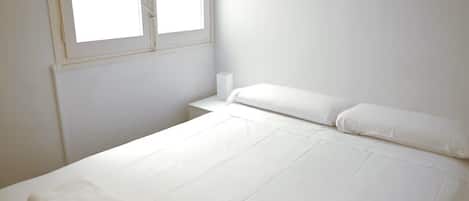 Un lit simple avec du linge de lit blanc, une fenêtre avec des stores et un mur blanc uni.