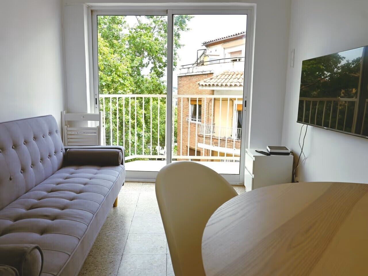 A room with a sofa, a chair, a table, and a balcony with a view of buildings and trees.