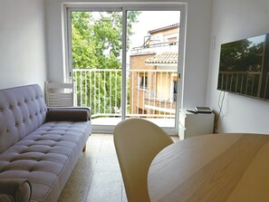 Ein Zimmer mit einer Couch, einem Sessel, einem Tisch und einem Balkon mit Blick auf Gebäude und Bäume.