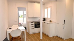 Une cuisine de taille réduite avec des meubles blancs, un coin repas comprenant une table et des chaises, et une fenêtre laissant entrer la lumière naturelle.