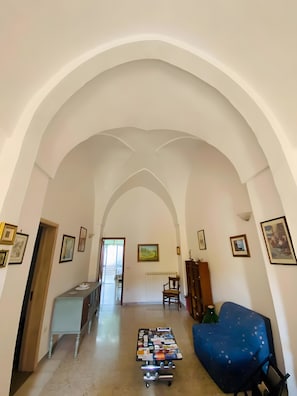 Un couloir doté de plafonds en arc de cercle, un coin salon avec un fauteuil recouvert de tissu, une table sur laquelle sont disposés divers objets, et des tableaux encadrés accrochés aux murs.