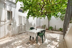 A patio with a table and chairs, a white wall, and a tree providing shade. - Salento Short Let Apartment: Spacious, Bright, Private Terrace, Garden, Air Conditioning (Copertino)