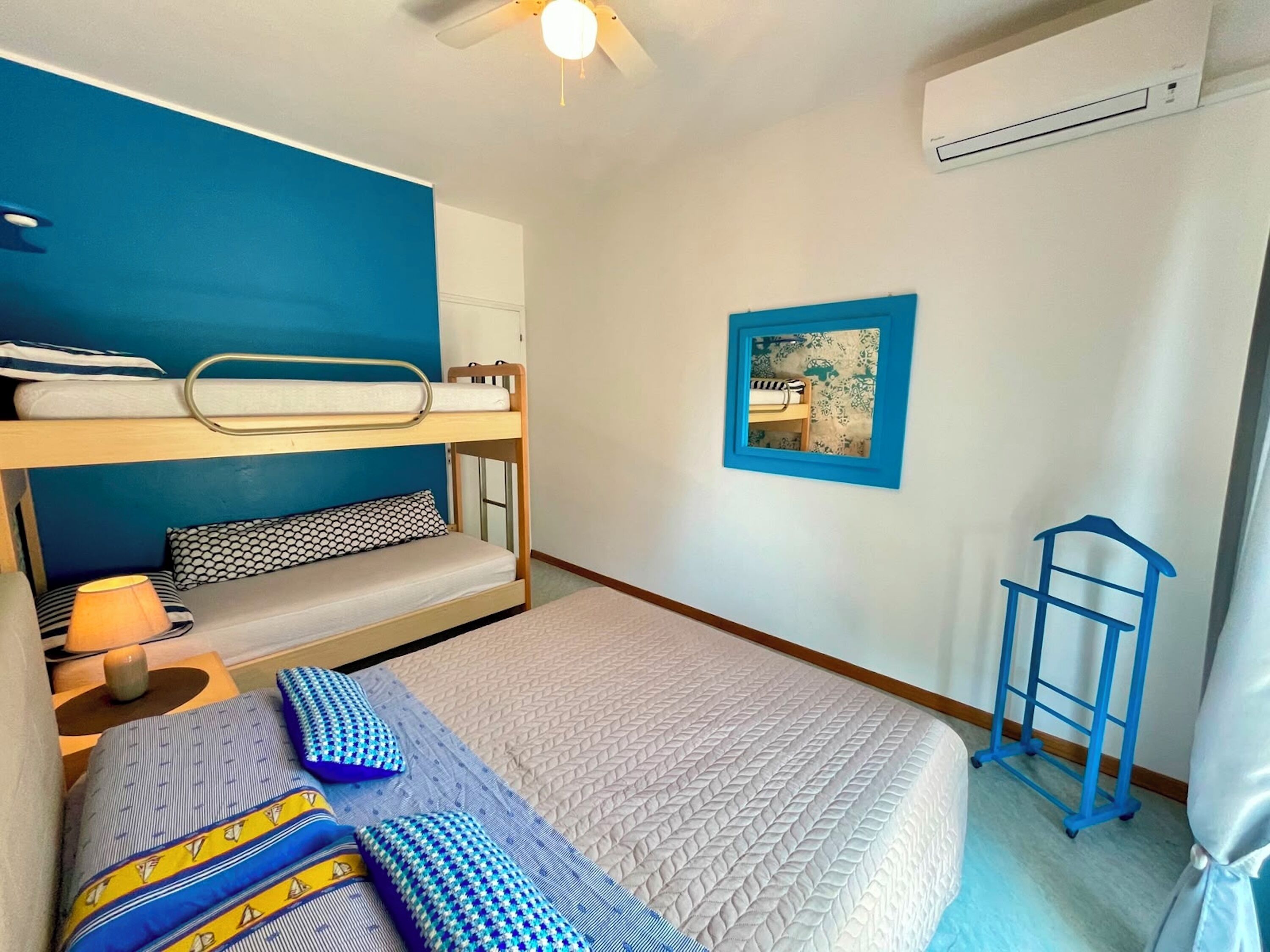 Een slaapkamer met stapelbedden, een plafondventilator, een klein tafeltje met lamp, een foto aan de muur en een blauwe stoel.