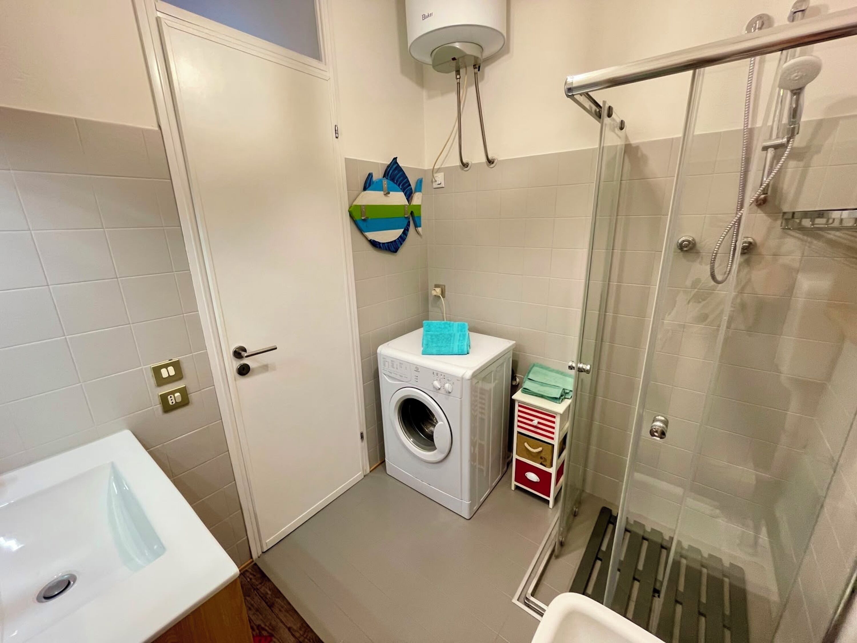 A compact bathroom with a washing machine, a glass shower enclosure, and a small storage unit.