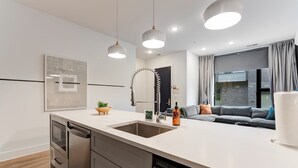 A modern kitchen with a central island, stainless steel appliances, pendant lighting, and a view to a living area with a gray sofa. - The Grove (Nashville)