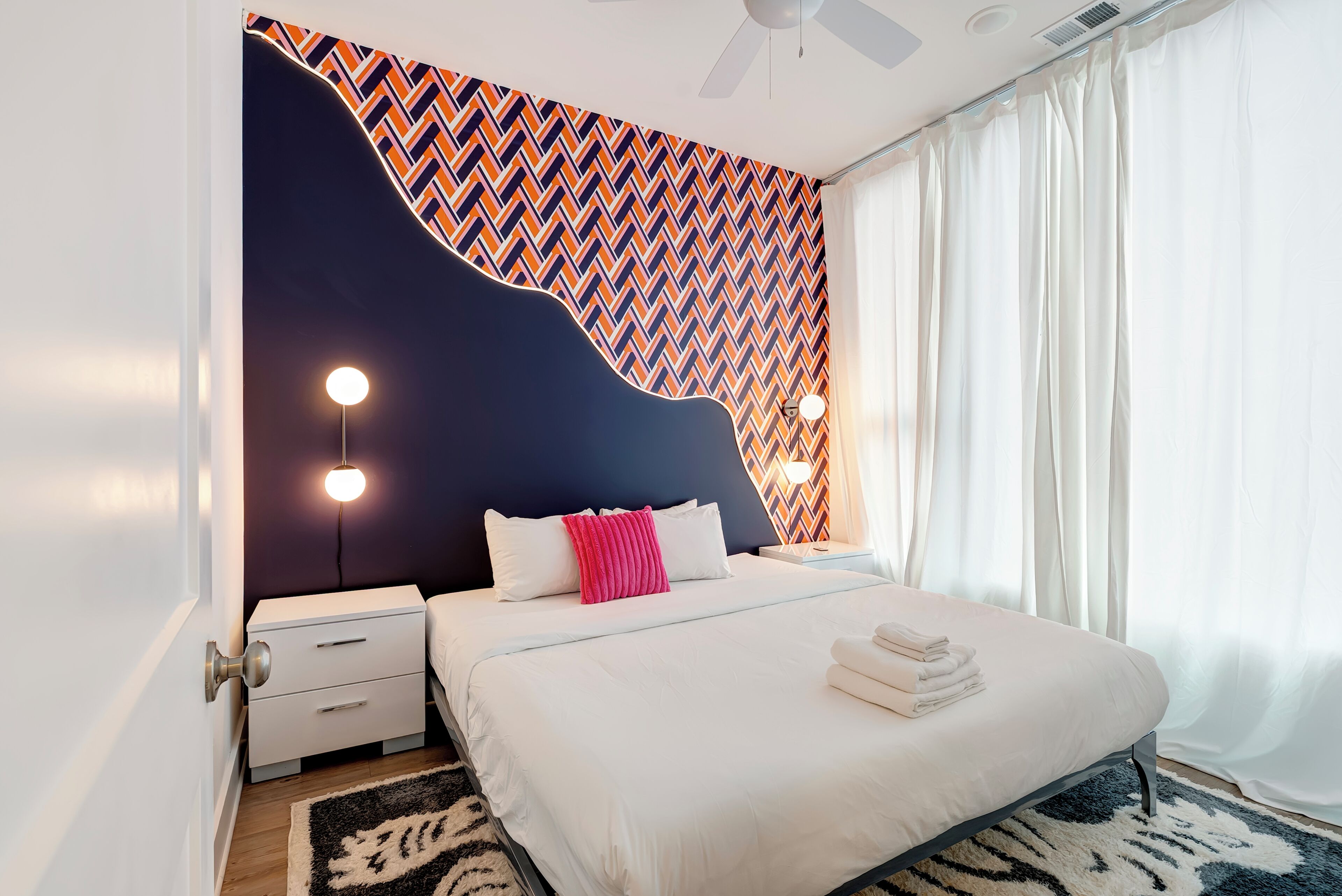 A modern bedroom with a unique geometric-patterned wall, a bed with white bedding, a nightstand, and a bedside lamp.