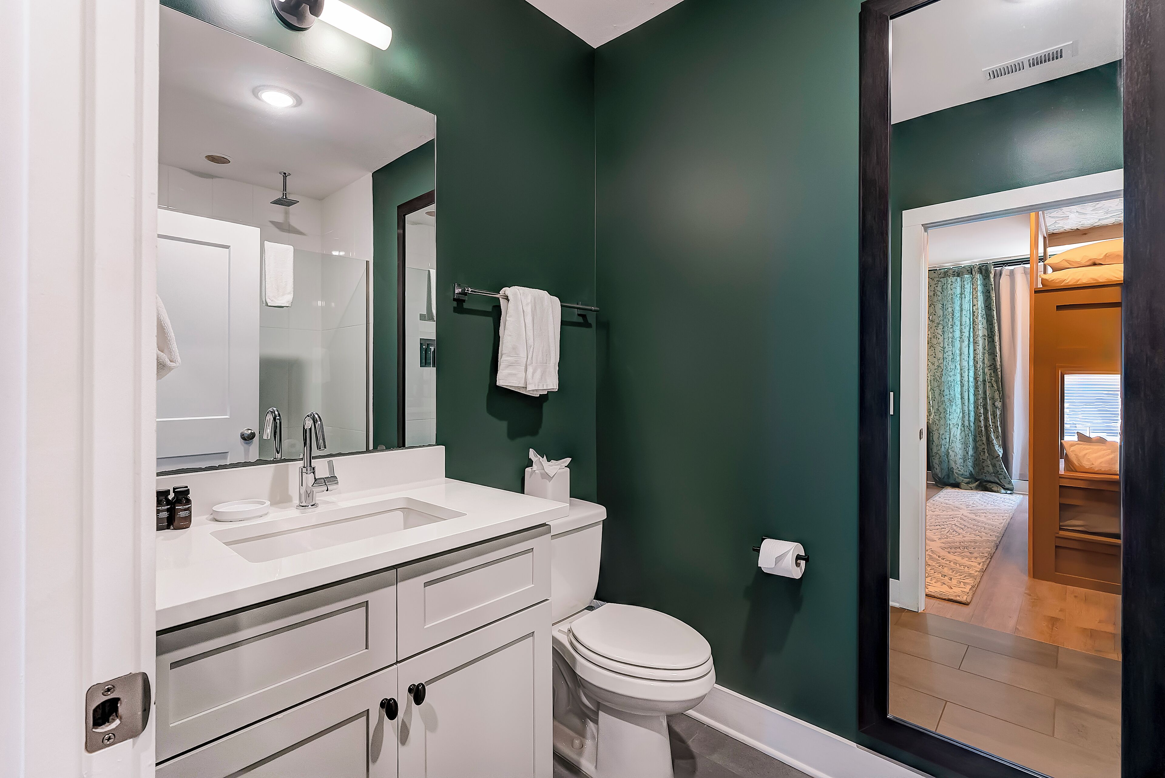 A bathroom with a white sink, mirror, and toilet, a towel rack, and a door leading to a bedroom with a bed and curtains.