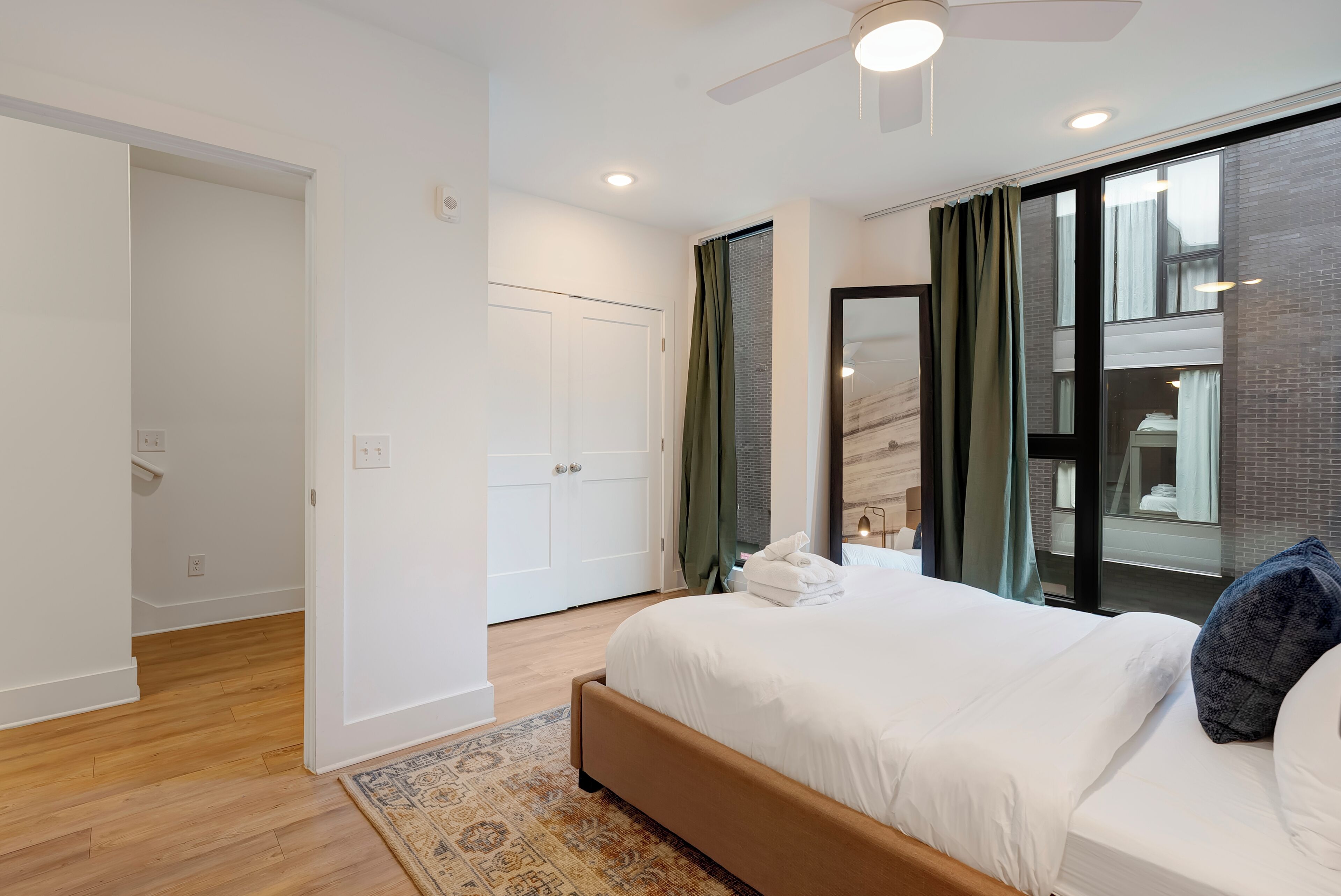 A bedroom with a bed, a ceiling fan, a window with curtains, and a view of a building outside.