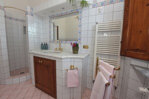 Un baño con ducha de azulejos blancos, lavamanos con detalles dorados, espejo con borde floral y toallero con toallas rosas.