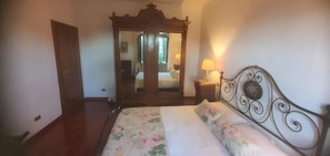 Un dormitorio con un armario de madera, una cama con ropa de cama floral, una mesita de noche con lámpara y un espejo sobre la cama.