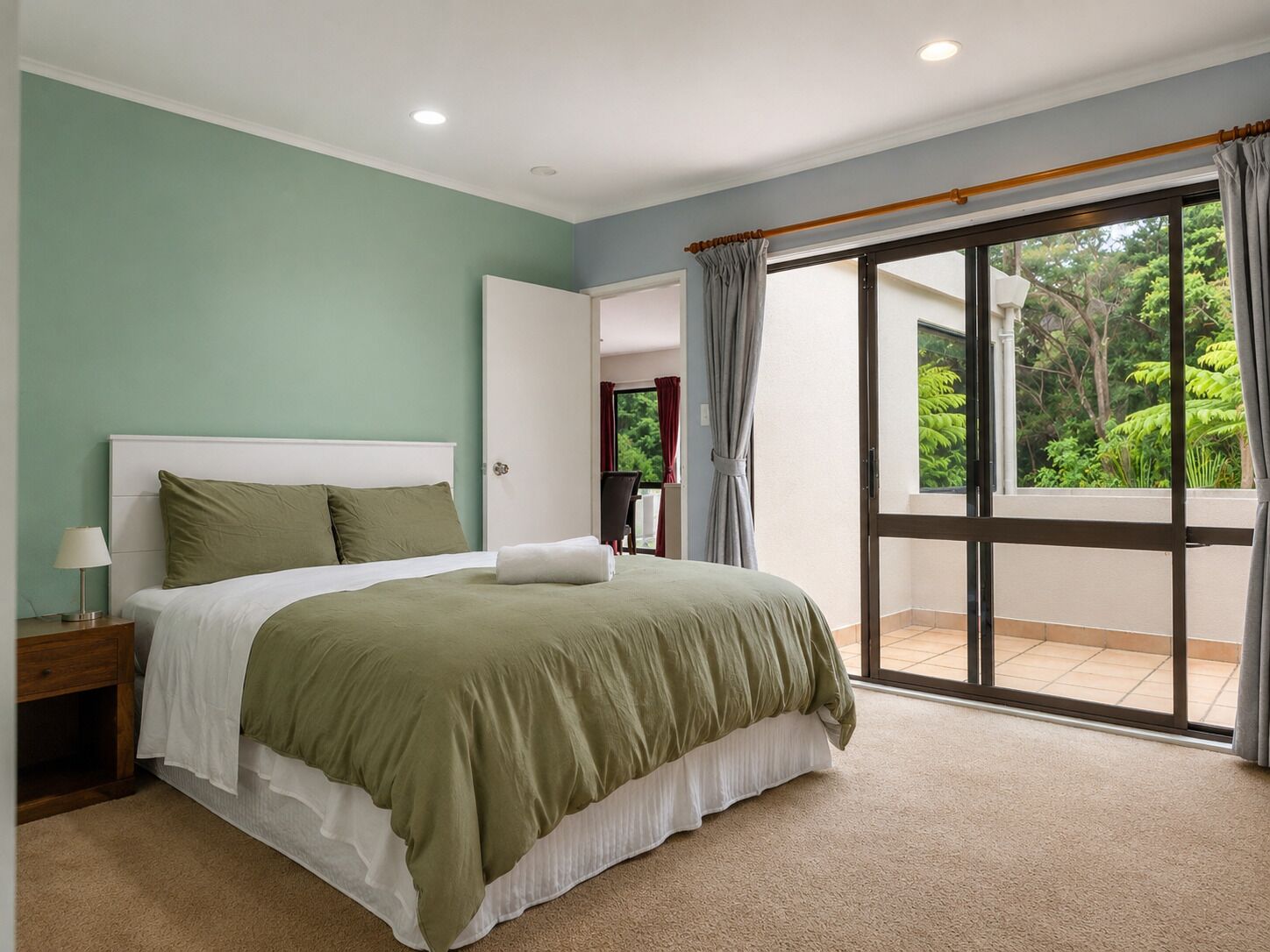 A bedroom with a bed, nightstand, and a view of the outdoors through a large window.