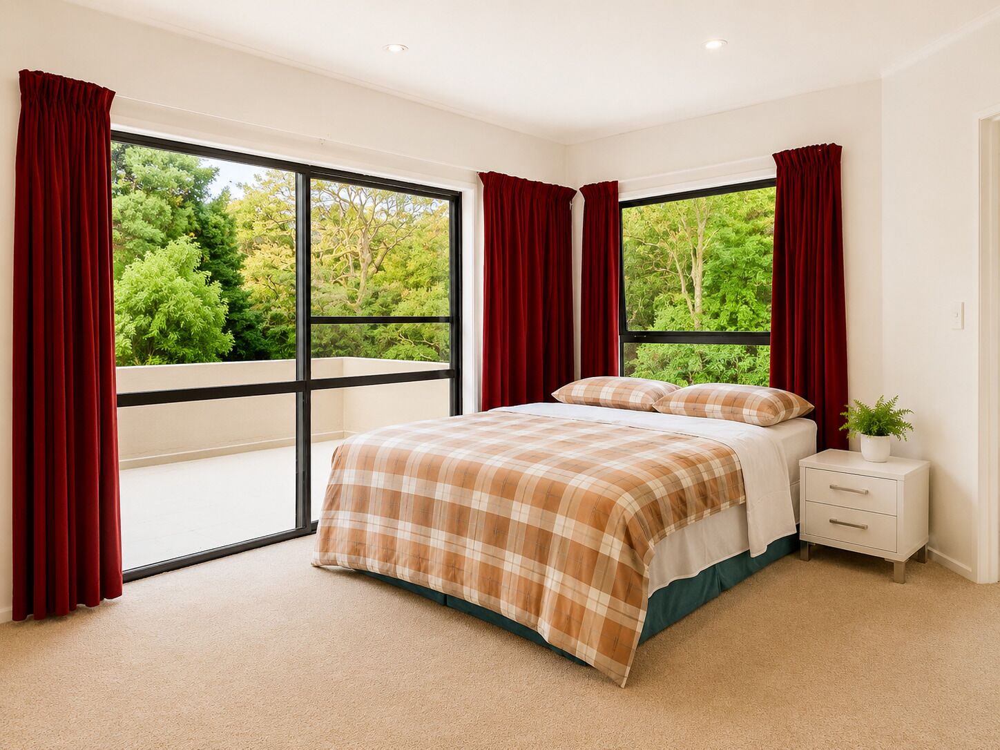 A bedroom with a large window, a bed with a plaid bedspread, a nightstand, and a potted plant.