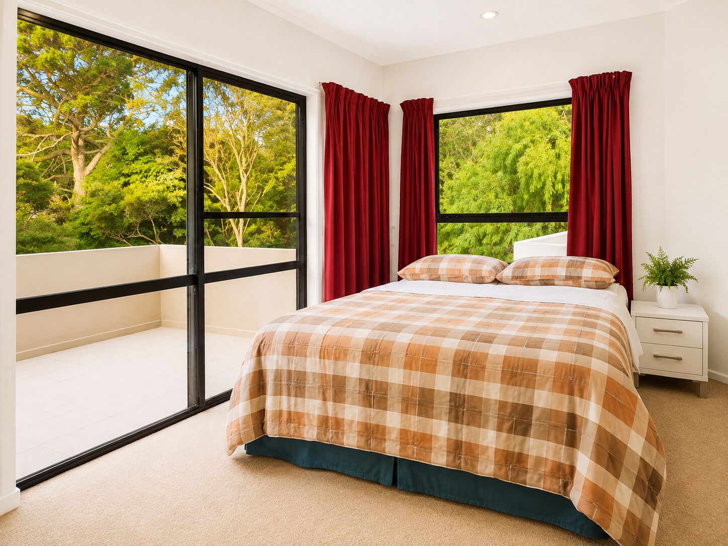 A bedroom with a large window, a bed with a plaid bedspread, red curtains, and a nightstand with a plant.