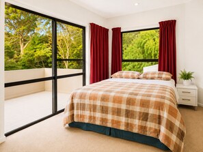 Un dormitorio con un ventanal grande, una cama con colcha escocesa, cortinas rojas y una mesita de noche con una planta.