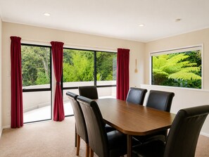 Un salón de conferencias con una gran mesa y cuatro sillas, ventanas amplias con cortinas rojas y vistas al exterior con vegetación.