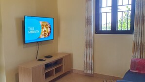 Una sala de estar con un televisor montado en la pared, un mueble bajo de madera, una ventana con cortinas y una puerta a la izquierda.