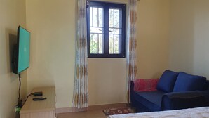 Una habitación con una cama, un sofá, un televisor montado en la pared, una ventana con cortinas y una mesita con un distribuidor de corriente.