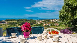 Une table dressée avec des tasses et des soucoupes blanches, un vase rose orné de fleurs et un saladier rempli de fraises, le tout offrant une vue sur un paysage côtier.