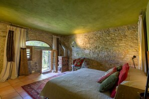 A bedroom with a stone wall, a bed with pillows, a chair, a wooden dresser, and a window with curtains. -  Chianti country retreat 10 pax (Radda in Chianti)