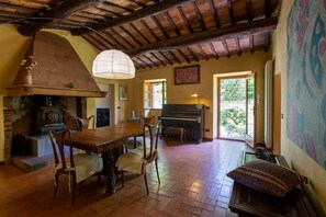 A rustic interior with a stone fireplace, wooden dining table, chairs, a piano, and a large window. -  Chianti country retreat 10 pax (Radda in Chianti)