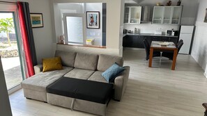 A modern living room with a sofa, a dining table with chairs, a kitchen area, and a view through a glass door. - Modern apartment with a terrace in the heart of town (Vila Franca Do Campo)