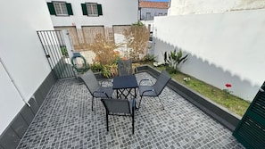 A patio with a table and chairs, a small garden area with plants and a rose, and a white wall with a window.