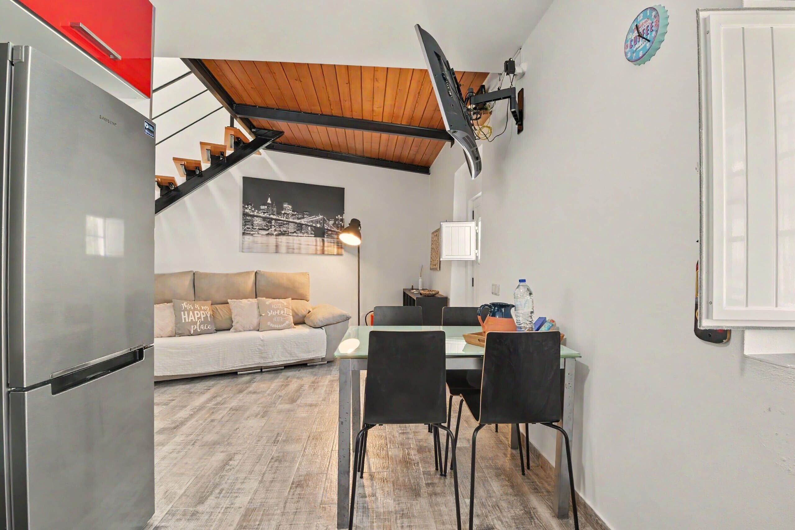 Een compacte keuken met een moderne koelkast, een eethoek met een tafel en stoelen, een bank met kussens, en een trap naar een hoger niveau.