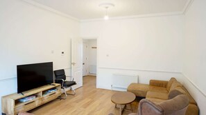 A living room with a flat-screen TV, a black chair, a brown sofa, a small round table, and a wooden TV stand. - Light Filled 2BDR Riverside Retreat Rotherhithe (London)