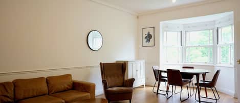 Un salon avec un canapé en cuir marron, une petite table ronde, un fauteuil marron, une table à manger avec quatre chaises et une grande fenêtre aux montants blancs.